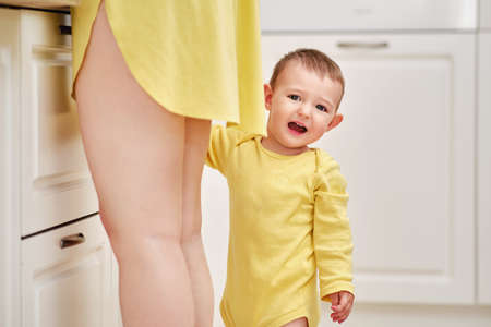 Unhappy toddler baby boy stands holding his mother legs in the kitchen, kid age one yearの写真素材