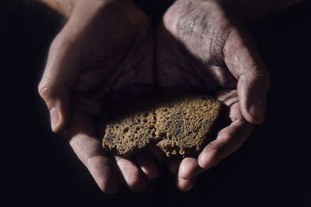 Hungry man holding bread on a black background, hands with food close-up. Dirty hands of a starving poor man on a dark backgroundの写真素材