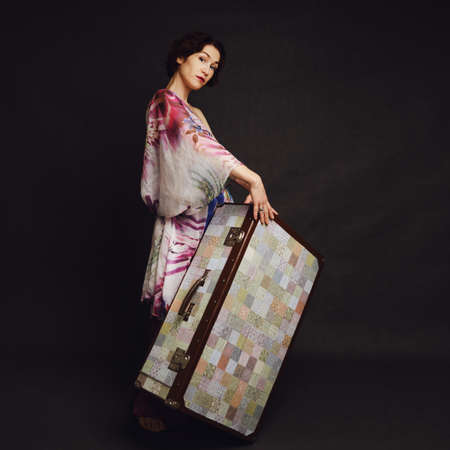 A happy woman in an Asian dress with a suitcase on a studio black background. Smiling woman with a bag on a dark backgroundの写真素材