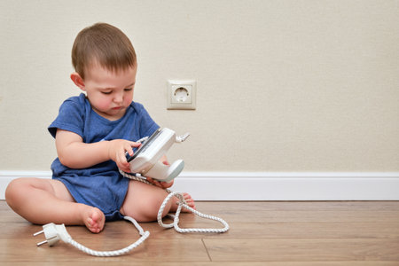 Toddler baby plays with a hot iron at the electrical outlet, copy space. Child boy holding a dangerous iron in his hands. Kid age one yearの写真素材