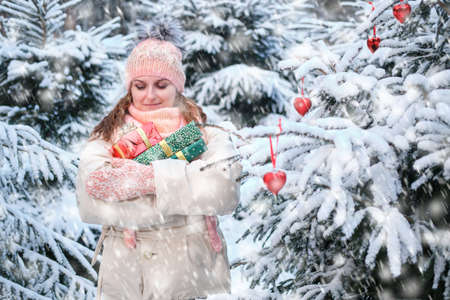 A happy woman in winter clothes stands with gifts at a Christmas tree in a snowy forest on New Year Eveの写真素材