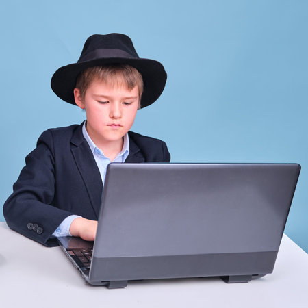 A boy in a school suit and a black hat at a computer during distance learning, copy space on a blue studio backgroundの写真素材
