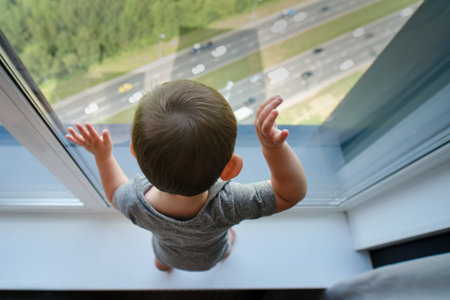 Toddler baby is standing by the window on the windowsill. Child at the window holding hands on the...