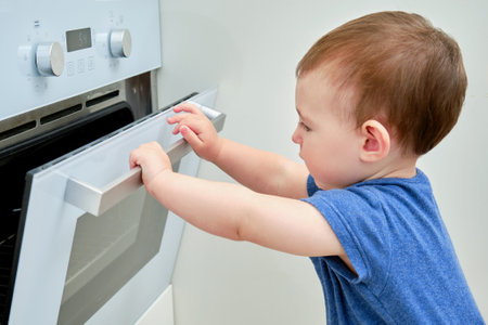 Toddler baby climbs into a hot electric oven. Child boy opens oven door in home kitchen. Kid age one yearの写真素材