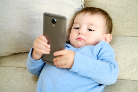 Doubting toddler baby boy is sitting with a phone on the sofa in the living room. Child with a smartphone in his hands on the bedの写真素材
