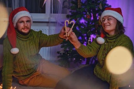 A man and a woman hold heart shaped caramel canes at the Christmas tree in the night light of a garland. Home comfort on New Year Eve in the evening living roomの写真素材