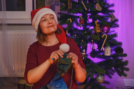 An adult woman knits clothes by the Christmas tree in the evening living room. Hobbies and needlework with wool yarn on New Year Eveの写真素材