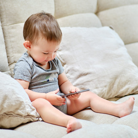 Happy toddler baby boy is sitting with a phone on the sofa in the living room. Child with a...