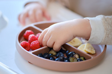 Toddler baby eats fruits and berries with his hand, table close-up. Child hands take food from a beige plate. Kid aged one year and two monthsの写真素材