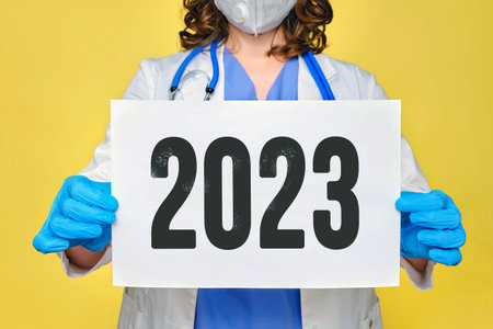 Woman doctor in a white uniform with a sheet A4 paper, text 2023 year on a yellow background