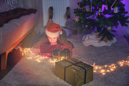 A lonely boy lies at the Christmas tree and looks at the gifts. Home family comfort on New Year Eve...