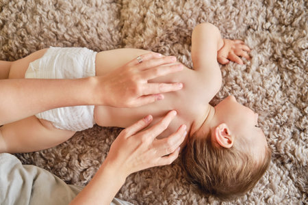 Woman mother doing back massage to happy toddler baby lying on beige bedspread. Mom masseur and smiling child boy. Kid aged one year and two monthsの写真素材