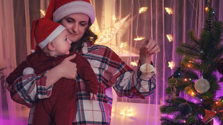 Baby boy toddler with his mother decorate the family Christmas tree with baubles on New Year Eve. Happy child in the hands of a woman in a santa hat decorates the Christmas treeの写真素材