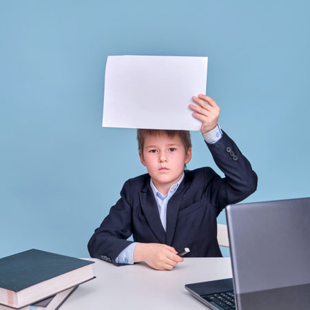 A boy in a school suit with a sheet of A4 paper at a computer during distance learning, copy space...