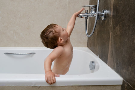 Happy toddler baby is playing in white bathtub with water faucet. Kid aged one year and two monthsの写真素材