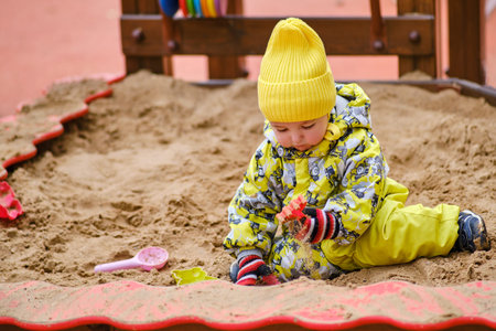 Happy toddler baby boy is playing in the sandbox. A child in warm clothes is playing with sand on...