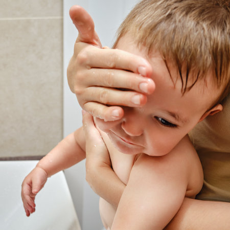 Woman mother washes face of sad toddler baby in sink with faucet. Mom helps with hygiene crying child boy. Kid aged one year and two monthsの写真素材
