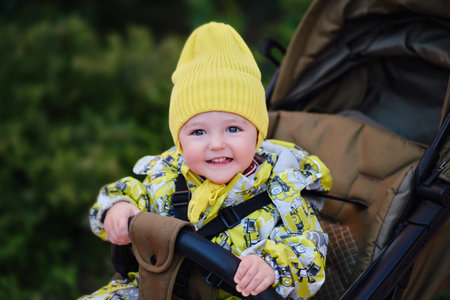 Happy toddler baby boy in yellow overalls sits in a stroller. A child in warm clothes against the...