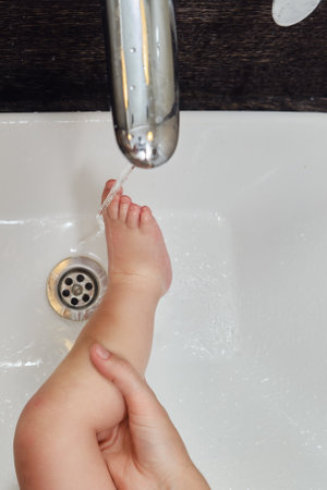 Woman mother washes legs of toddler baby in sink with faucet. Mom helps with hygiene foot of child boy. Kid aged one year and two monthsの写真素材