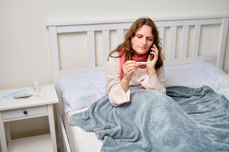 Sick woman calls the doctor on the phone with a thermometer in her hand, fever. Adult ill woman wrapped in a red scarf sits on a home bed in a white bedroom, female age 35の写真素材