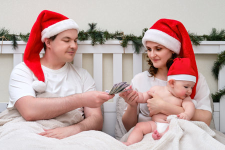 Husband gives money dollars to wife with child on bed decorated for christmas and new year, family couple in home bedroomの写真素材