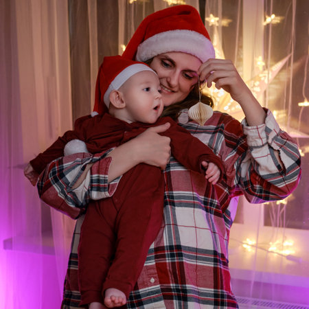 Baby boy toddler with his mom decorate the family Christmas tree with baubles on New Year Eve. A child in the hands of a woman in a santa hat decorates the Christmas treeの写真素材