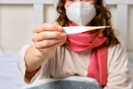 A sick woman is sitting on a bed in a medical face mask and a thermometer in her hands. Adult ill...