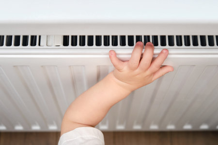 Toddler baby holding on to the radiator, child hand on the heating system close-up. White radiator for heating the home room and children's handの写真素材