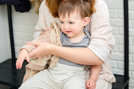 A mother puts a hoodie jacket on a toddler baby sitting in the home hallway. Woman mom dressing warm clothes on child for winter walk in cold weather. Kid aged one year and three monthsの写真素材