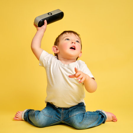 Toddler baby plays with a wireless music speaker on a studio yellow background. Happy child in a white t-shirt and blue jeans listens to music in an audio speaker. Kid aged one year four monthsの写真素材