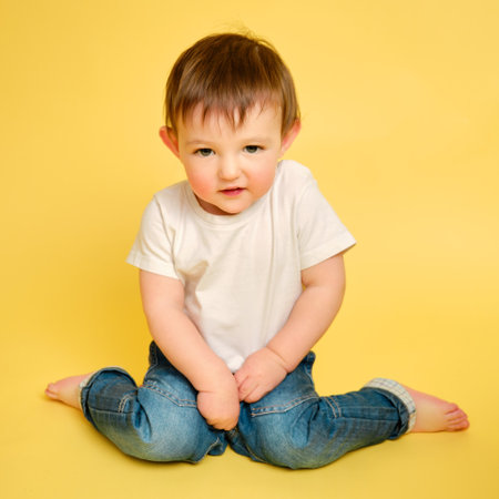 Portrait of a shy toddler baby in full length on a studio yellow background. Embarrassed child sitting on the floor in a white t-shirt and blue jeans. Kid aged one year and four monthsの写真素材