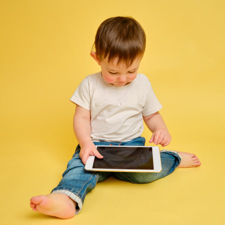 Toddler baby with a digital tablet on a studio yellow background. Happy child playing with a tablet in a white t-shirt and blue jeans. Kid aged one year and four monthsの写真素材