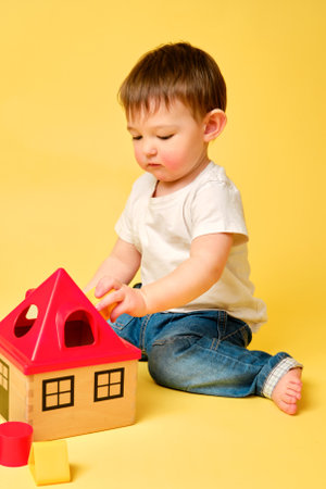 Toddler baby is playing sorter logic educational games with geometric shapes on a studio yellow background. Happy child play with educational toy house, learning logic. Kid aged one year four monthsの写真素材