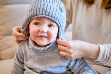 Child is dressed in warm winter clothes for a walk outside. Woman mother puts clothes on toddler baby boy for cold weather. Kid age one year five months in the home living roomの写真素材