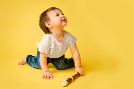 Toddler baby with a flute wind musical instrument on a studio yellow background. A happy child...