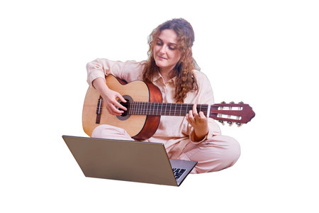 Young woman plays the guitar while sitting on the bed with a cat, online music lessons, isolated on a white background. Woman with a musical instrument in her hands in the home bedroom with laptopの写真素材