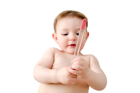 Child cleans his mouth with a toothbrush in his hands, isolated on a white background. Happy toddler baby learns to brush her teeth while lying on her home bed. Kid aged one yearの写真素材