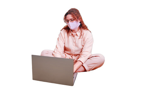 A nice woman in a medical mask is sitting on a home bed and remotely working on a laptop, isolated...