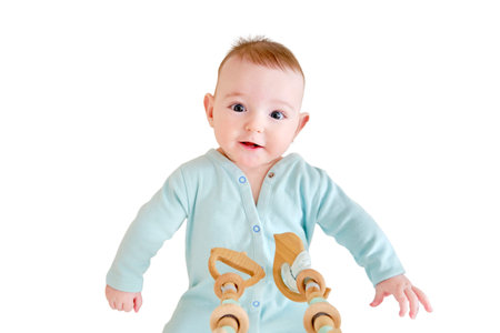 Happy infant baby is playing with wooden hanging toys lying in bed, isolated on a white background. Smiling funny child in turquoise clothes in a crib, aged six monthsの写真素材