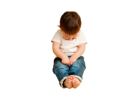 Full length portrait of an embarrassed toddler baby on a studio isolated on a white background. Shy child sitting on the floor in a white t-shirt and blue jeans. Kid aged one year and four monthsの写真素材