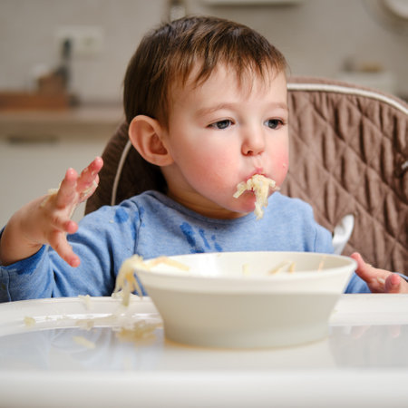 A funny child is eating a grated apple with his mouth full while sitting on a kitchen chair. Hungry baby boy shoves food in his mouth, humor. Kid aged one year four monthsの写真素材