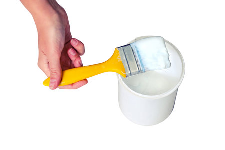 A woman's hand dips a yellow brush into a jar of white paint, isolated on a white background. Painting of walls and other surfaces during repair with white enamel.の写真素材