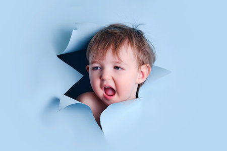 A yawning baby in a hole on a paper blue background. Torn child's head studio background, copy space. Kid aged one year six monthsの写真素材