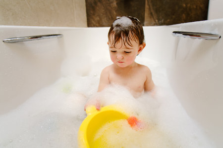 A happy child is playing with toys while sitting in a white homemade bathtub. Little toddler baby boy bathes in a bubble bath. Kid aged one year six monthsの写真素材