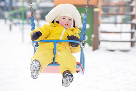 Happy toddler baby rides on a swing on a winter playground. Baby boy in yellow snowsuit swinging in kindergarten. Kid age one year eight monthsの写真素材