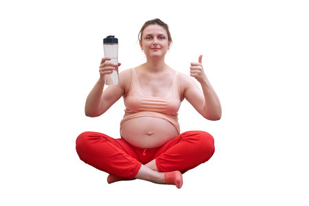 Fitness during pregnancy and the benefits of clean water, isolated on a white background. Pregnant woman at sports training at home shows the thumbs up with a bottle of water in her handsの写真素材