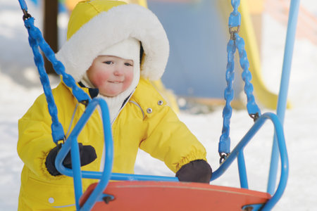 Happy toddler baby himself climbs on the swing in the winter playground. Baby boy in yellow snowsuit swinging in kindergarten. Kid age one year eight monthsの写真素材