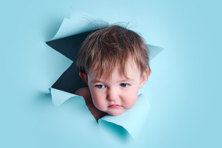 Sad baby in a hole on a paper blue background. Torn child's head studio background, copy space. Kid aged one year six monthsの写真素材