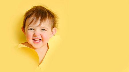 Happy baby in a hole on a paper yellow background. Torn child's head studio background, copy space. Kid aged one year six monthsの写真素材