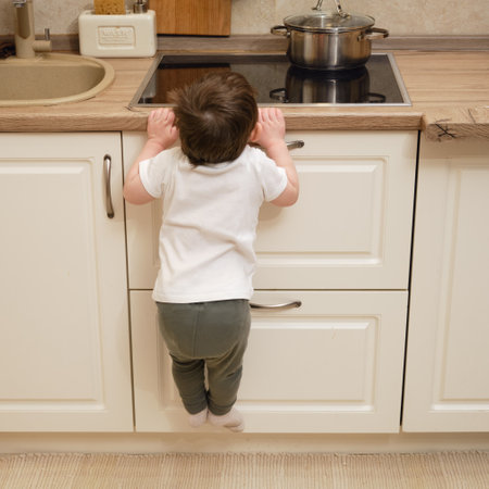Toddler baby climbs on a hot electric stove in the home kitchen. A small child touches the surface of the stove with his hand at the risk of getting burned. Kid aged one year eight monthsの写真素材